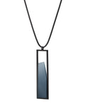 side view of The Lucia necklace features a sleek rectangular pendant that emphasizes form and material. A cast resin element rests within a black frame, creating a sense of depth and quiet luminosity. The elongated shape draws the eye, offering a refined balance between structure and softness. Minimal and striking, the Lucia necklace adds a sculptural accent that complements both everyday and statement looks.