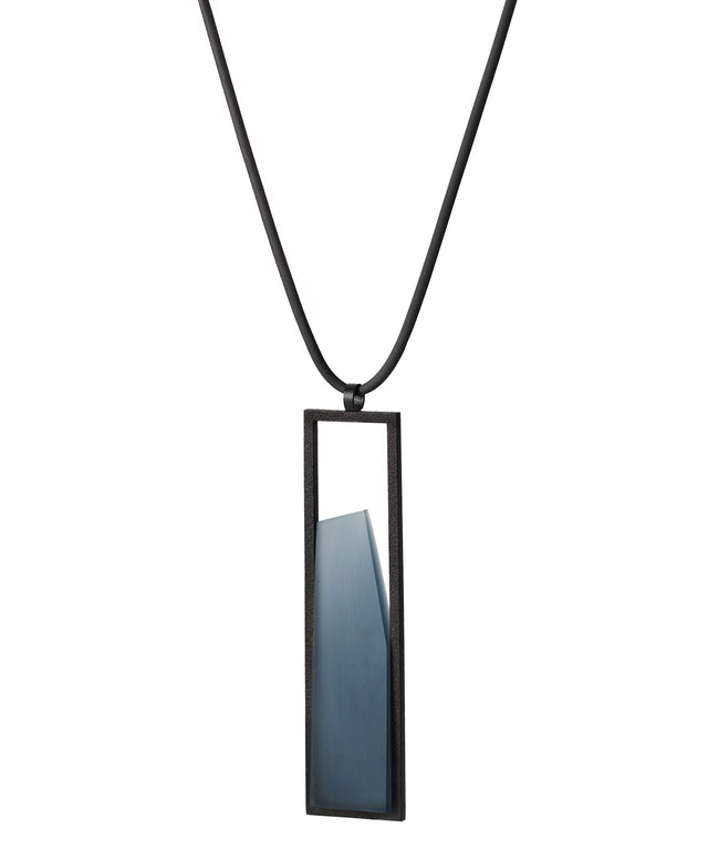 side view of The Lucia necklace features a sleek rectangular pendant that emphasizes form and material. A cast resin element rests within a black frame, creating a sense of depth and quiet luminosity. The elongated shape draws the eye, offering a refined balance between structure and softness. Minimal and striking, the Lucia necklace adds a sculptural accent that complements both everyday and statement looks.