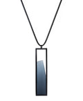 The Lucia necklace features a sleek rectangular pendant that emphasizes form and material. A cast resin element rests within a black frame, creating a sense of depth and quiet luminosity. The elongated shape draws the eye, offering a refined balance between structure and softness. Minimal and striking, the Lucia necklace adds a sculptural accent that complements both everyday and statement looks.