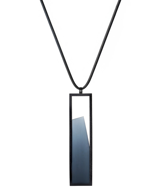 The Lucia necklace features a sleek rectangular pendant that emphasizes form and material. A cast resin element rests within a black frame, creating a sense of depth and quiet luminosity. The elongated shape draws the eye, offering a refined balance between structure and softness. Minimal and striking, the Lucia necklace adds a sculptural accent that complements both everyday and statement looks.