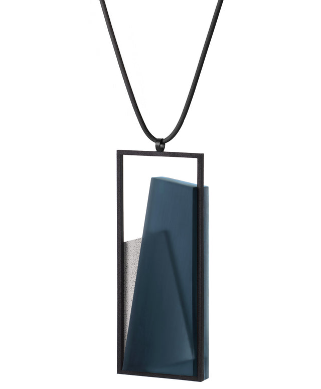 Side view of The Naia necklace in Cerulean blue embodies the feeling of wearing a work of art. Within its black rectangular frame, layers of resin and concrete are composed with sculptural precision, creating depth and quiet movement. The graduated tone of the resin shifts gently with the light, giving the piece a quiet, sculptural presence. Balanced and distinctive, the Naia necklace transforms simple geometry into a refined expression of wearable art.