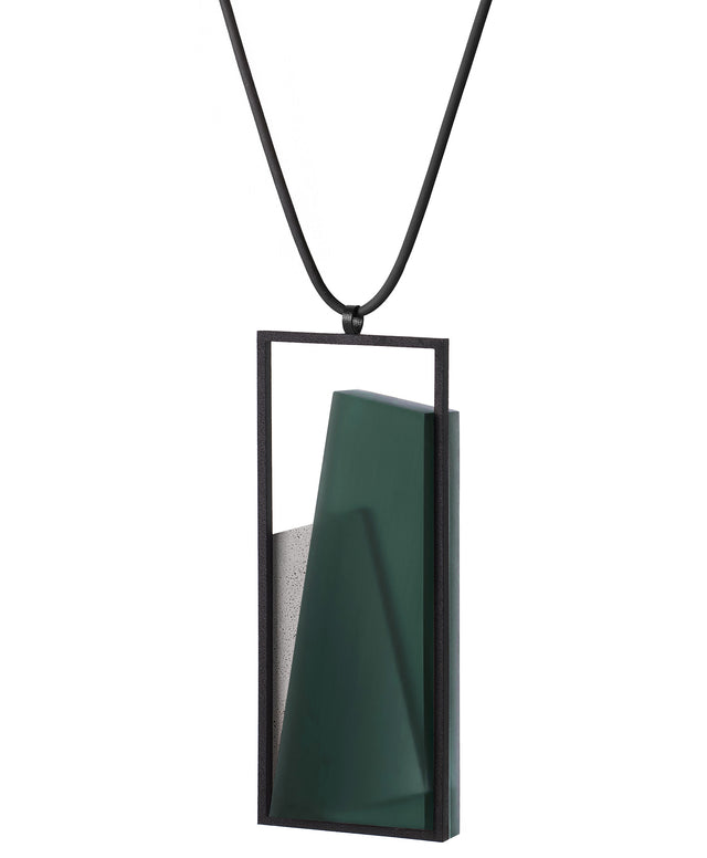 side view of the The Naia necklace in Cypress Green embodies the feeling of wearing a work of art. Within its black rectangular frame, layers of resin and concrete are composed with sculptural precision, creating depth and quiet movement. The graduated tone of the resin shifts gently with the light, giving the piece a quiet, sculptural presence. Balanced and distinctive, the Naia necklace transforms simple geometry into a refined expression of wearable art.
