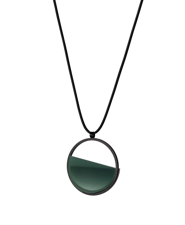 side version of the The Vestra Small necklace in cypress green resin captures the same sense of balance and quiet strength in a more understated form that the larger version. Named from the Latin vestra, meaning “yours,” it reflects a personal connection between light, material, and wearer. The smaller circular frame holds a smooth resin plane with a cast concrete inset that reveals gentle depth and tone, creating a soft glow close to the body.