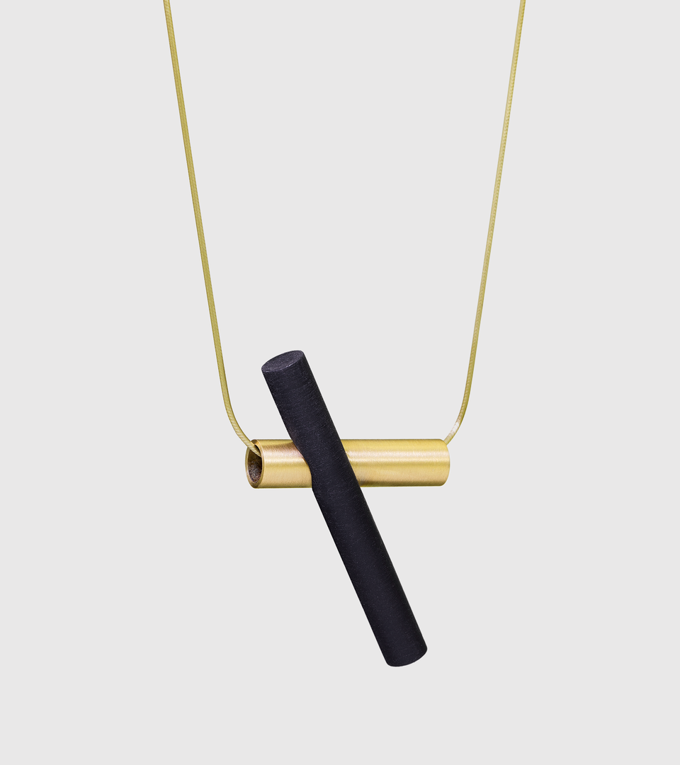 Suspended from a 14k gold snake-chain a black-tinted concrete cylinder cantilevers a brushed tube in solid 14k gold. 